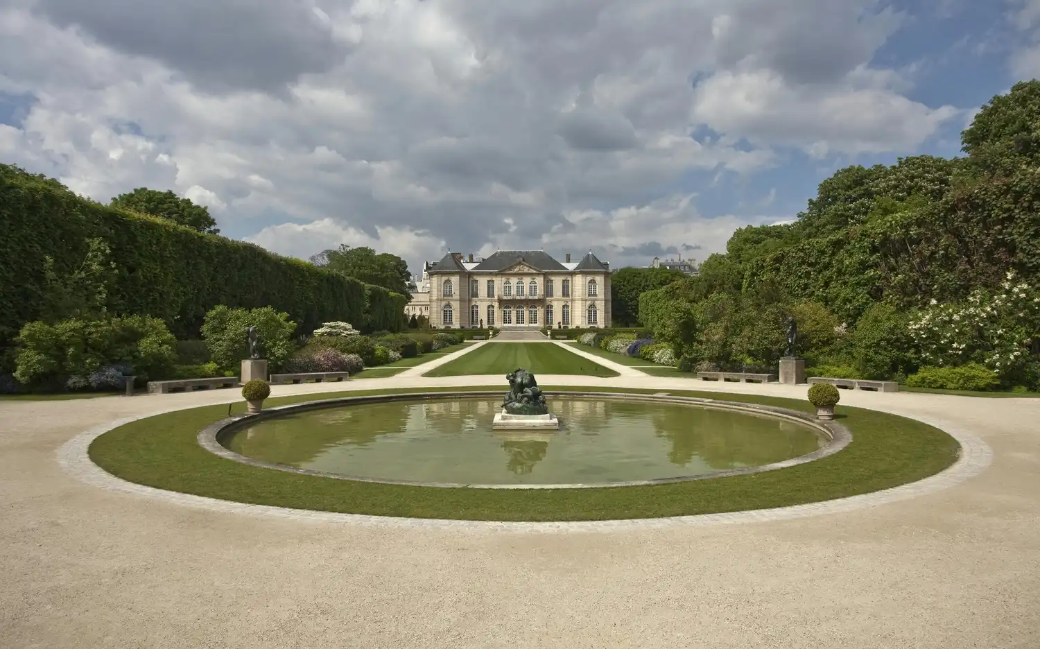 Грандиозный особняк виден через ухоженный сад с центральным прудом со статуей, окруженной статуями и подстриженными кустами.