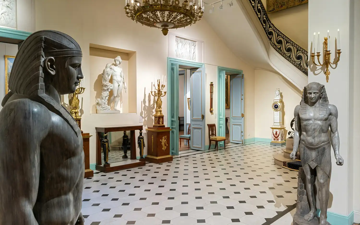Intérieur du musée avec des statues égyptiennes, des sculptures classiques, des lustres ornés et des meubles anciens dans une salle carrelée avec des portes bleu pâle.