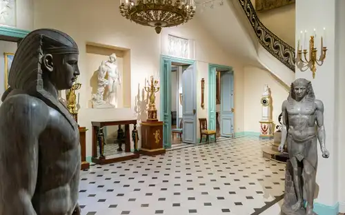 Intérieur du musée avec des statues égyptiennes, des sculptures classiques, des lustres ornés et des meubles anciens dans une salle carrelée avec des portes bleu pâle.