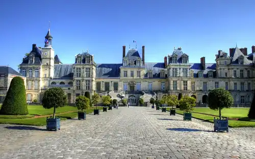 Historyczna rezydencja królewska Château de Fontainebleau Château de Fontainebleau z wieżami, brukowaną ścieżką i zadbanymi ogrodami pod bezchmurnym niebem.
