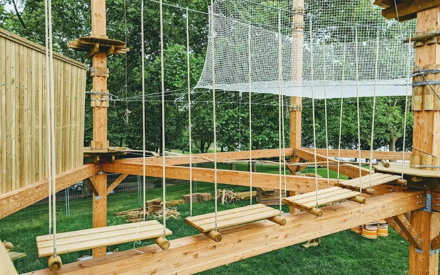 Wooden rope bridge and net structure in an outdoor adventure park, surrounded by green trees and grass. Wooden rope bridge and net structure in an outdoor adventure park, surrounded by green trees and grass.