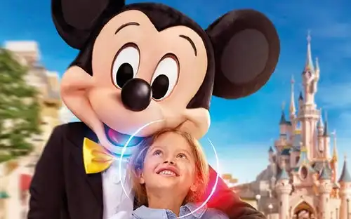 Zaczarowane bramy Disneyland® w Paryżu Dziecko uśmiecha się do Myszki Miki na tle bajkowego zamku i kolorowych budynków w Disneyland® w Paryżu.
