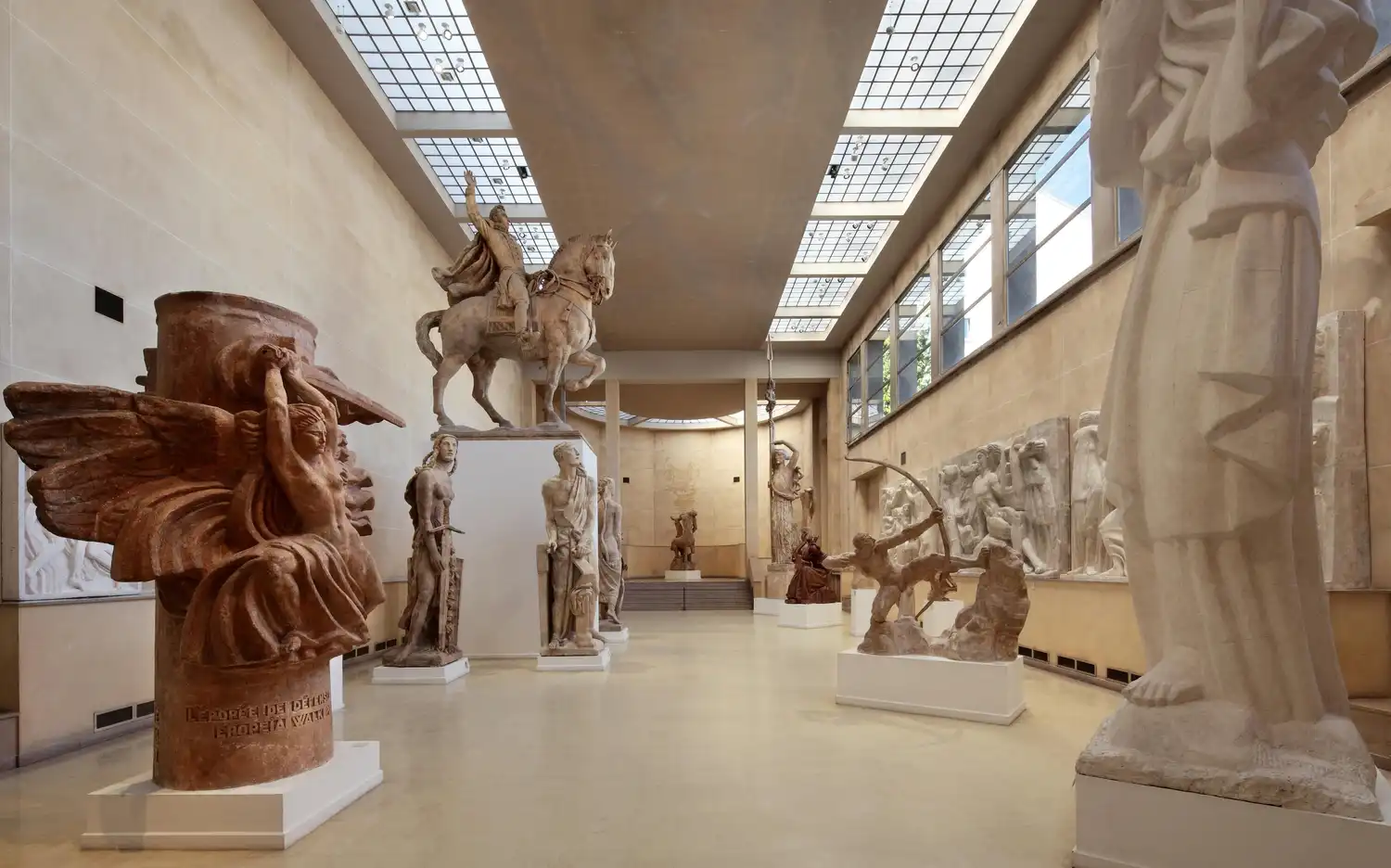 Une galerie de musée avec plusieurs grandes sculptures, dont un cavalier, une figure ailée et plusieurs statues humaines, éclairées par des puits de lumière. Une galerie de musée avec plusieurs grandes sculptures, dont un cavalier, une figure ailée et plusieurs statues humaines, éclairées par des puits de lumière.