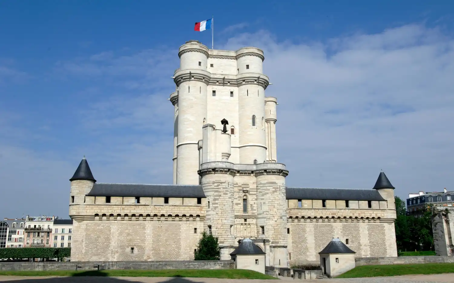 Wysoki kamienny Château de Vincennes z wieżyczkami i francuską flagą pod błękitnym niebem z rzadkimi chmurami.