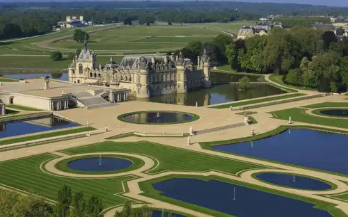 Vue aérienne d'un château orné, entouré de pelouses entretenues et de bassins avec fontaines, situé dans un vaste paysage verdoyant.