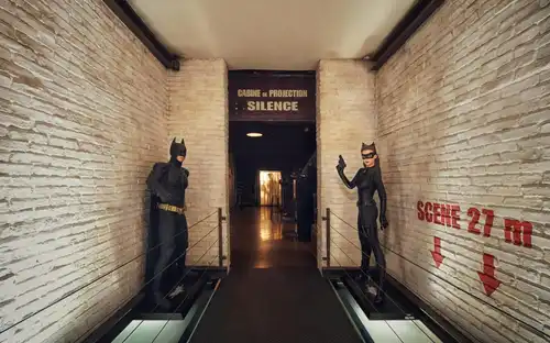 Couloir avec des statues de Batman et de Catwoman de chaque côté menant à l'entrée d'une salle de projection marquée "SILENCE".