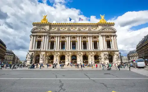 Un grand bâtiment néoclassique orné de statues dorées, des gens marchant devant, et un drapeau français flottant au sommet sous un ciel bleu.