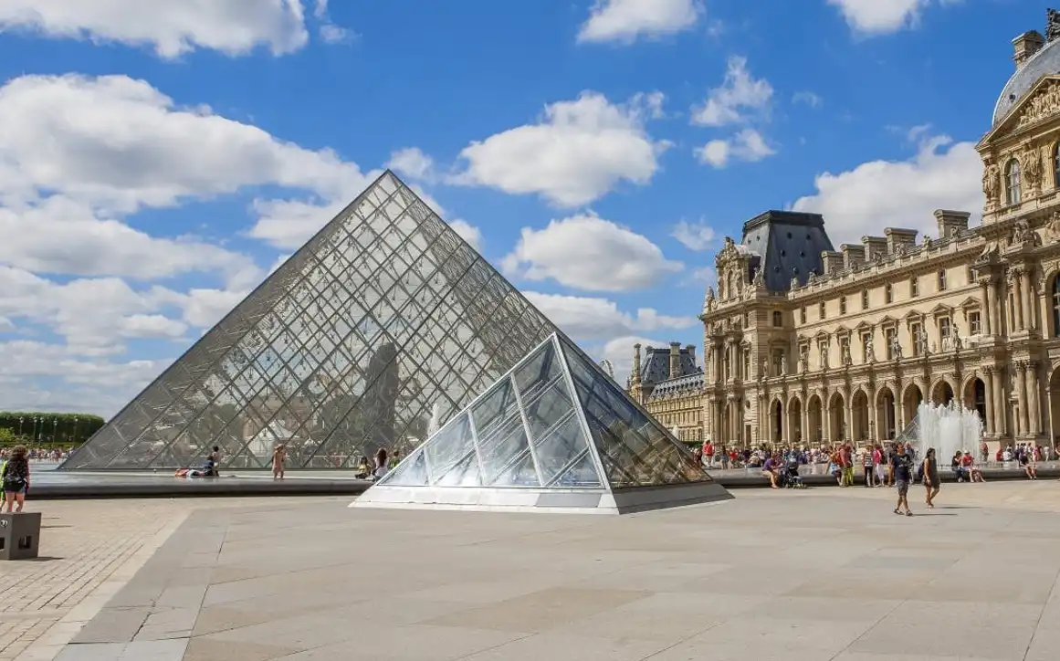 Glass pyramids at the Louvre Museum with people walking and resting around, historic building and blue sky with clouds in the background.