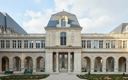 Historyczny dziedziniec Muzeum Carnavalet Zabytkowy budynek z łukowatymi oknami i zielony dziedziniec z ławkami pod błękitnym niebem w Muzeum Carnavalet.