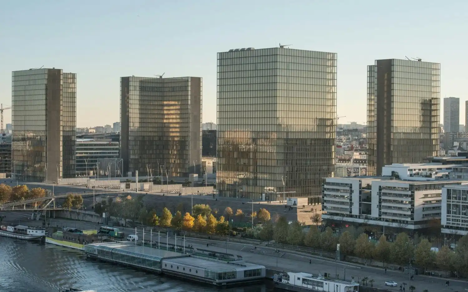 Bibliothèque nationale de France François-Mitterrand obejmuje cztery nowoczesne szklane budynki w kształcie otwartych książek stojące na brzegu rzeki pośród bujnych drzew i miejskiej zabudowy pod bezchmurnym niebem.