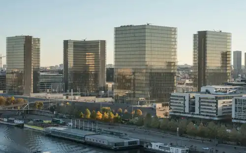 La Biblioteca nazionale di Francia François-Mitterrand presenta quattro moderni edifici in vetro a forma di libri aperti che sorgono sulla riva di un fiume con alberi rigogliosi e strutture urbane sotto un cielo sereno.